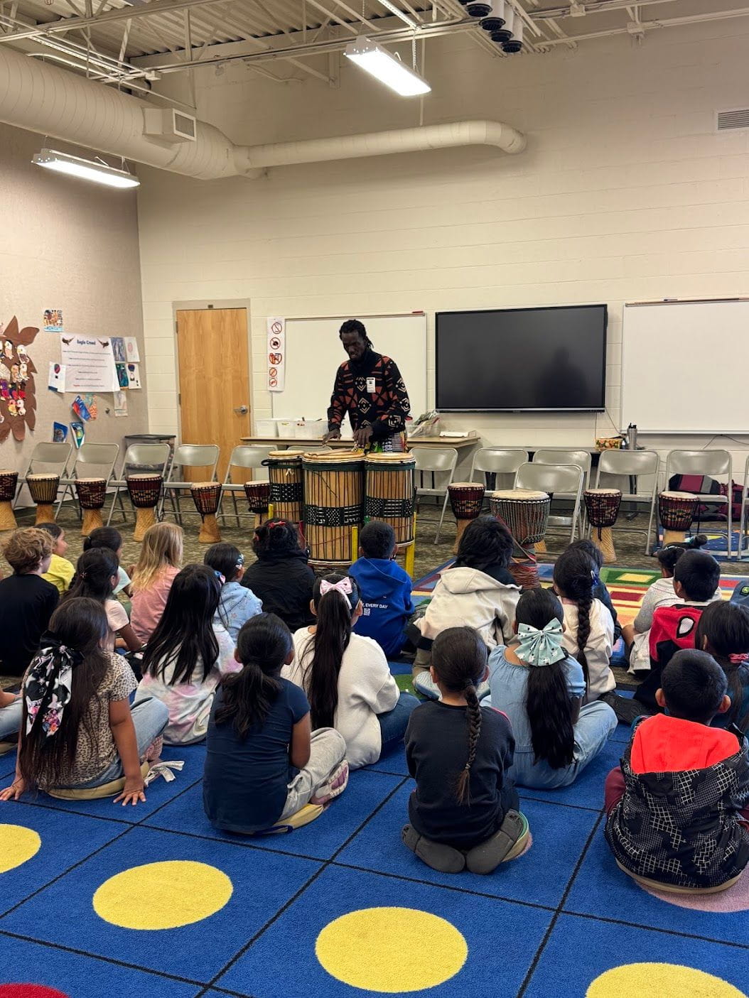 Classroom with kids sitting, watching a man play drums.