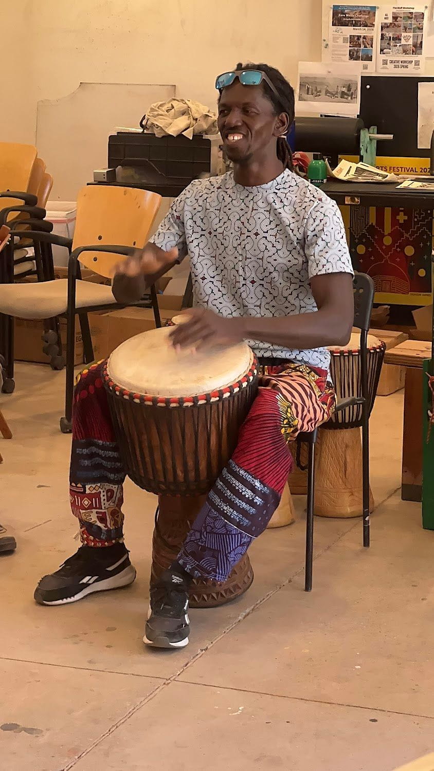 Man playing a djembe drum while seated, wearing colorful pants.