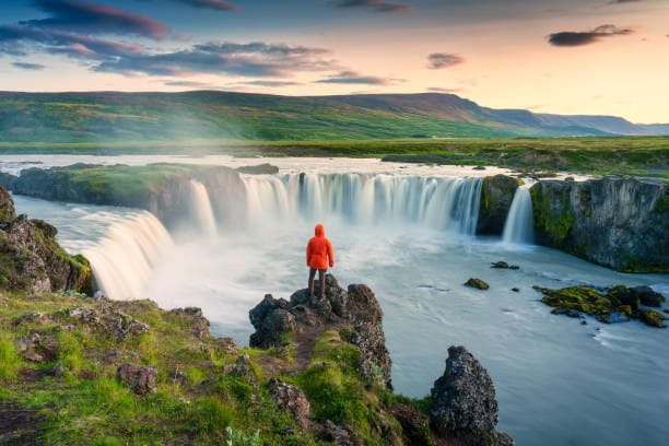 Godafoss waterfall flowing with colorful sunset sky and male tourist standing on cliff in summer at Iceland Majestic landscape of Godafoss waterfall flowing with colorful sunset sky and male tourist standing at the cliff on Skjalfandafljot river in summer at Northern Iceland iceland tourist stock pictures, royalty-free photos & images