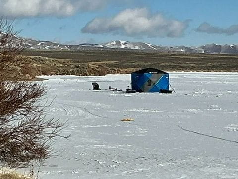 An ice fishing shelter sits on a frozen section of Lake John with some clouds in a blue sky and snowy mountains in the distance.