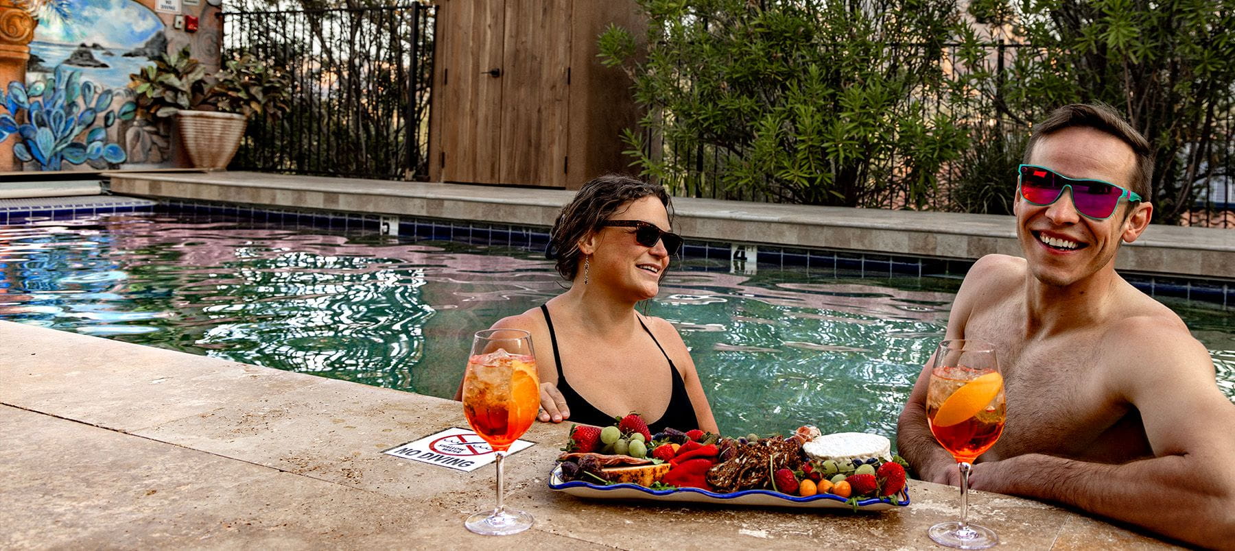 A smiling woman and man relax in a pool with a plate of fruit and drinks.