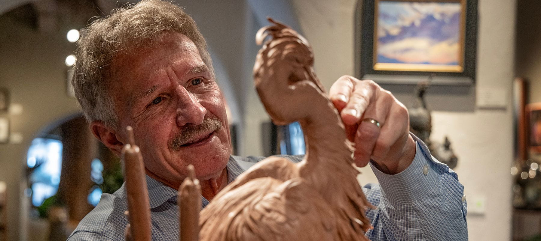 A smiling man uses a handheld tool to shape a sculpture of a bird.