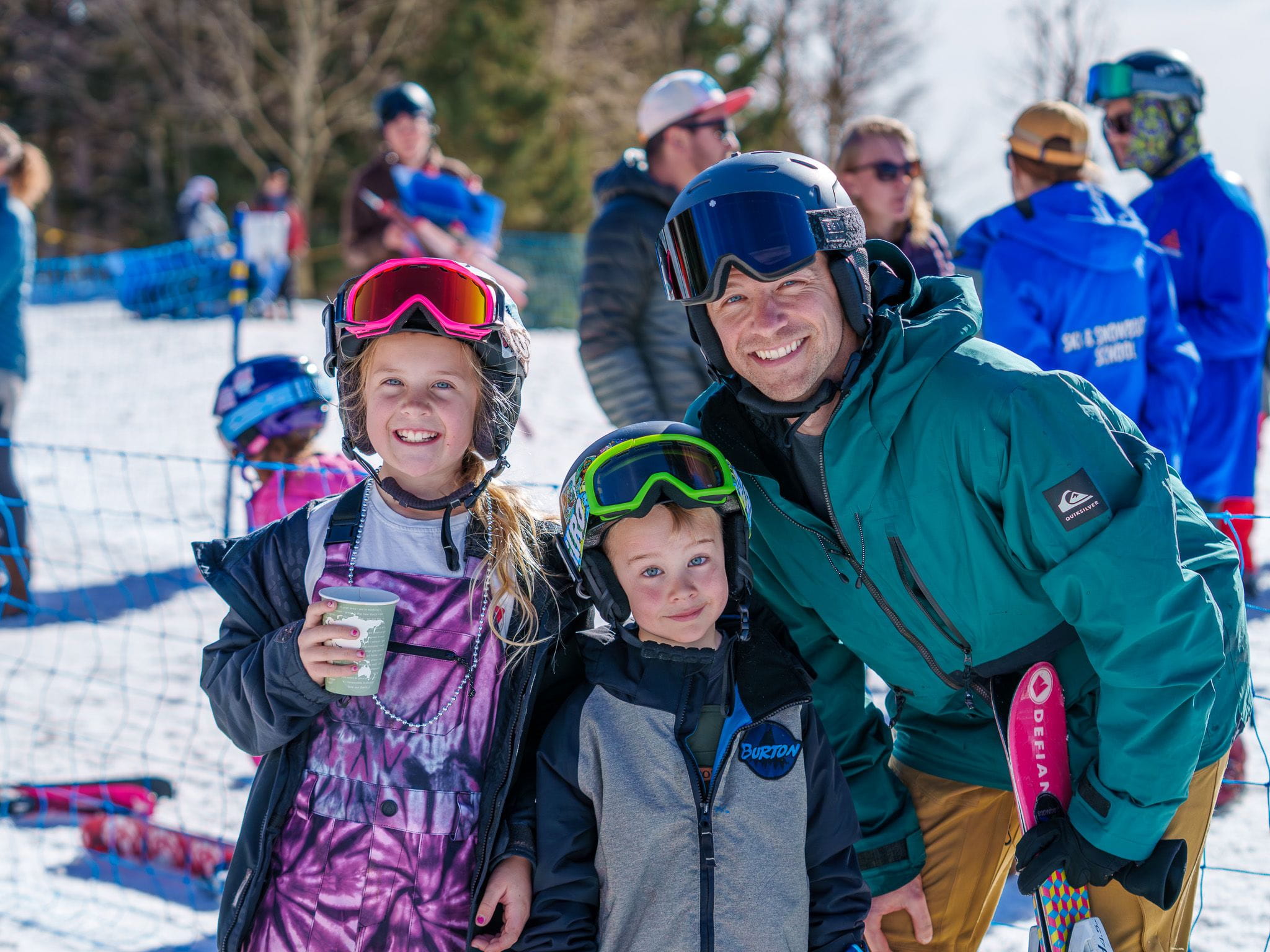 Two smiling children with a father all wearing ski gear in the snow
