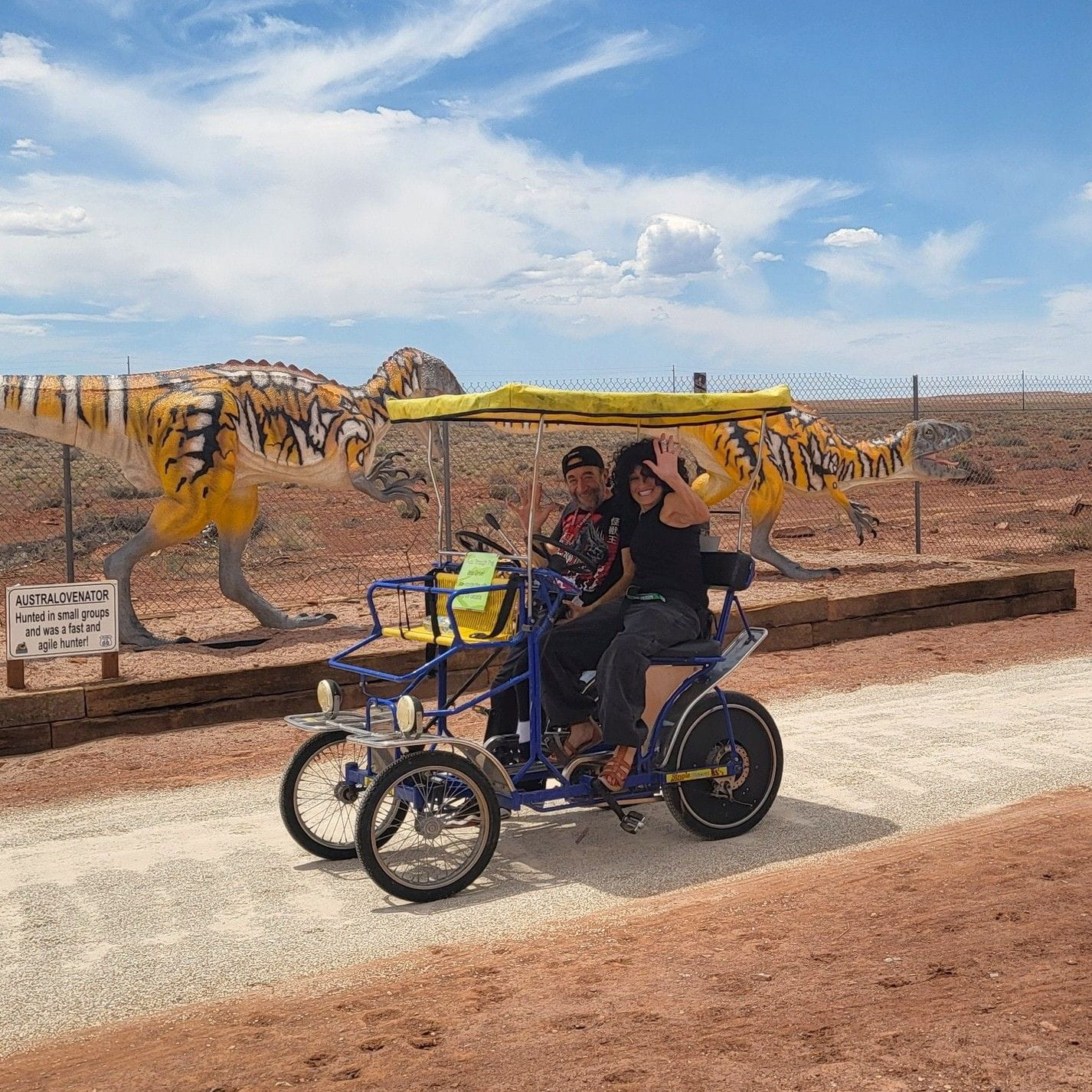 couple on a bicycle in the desert in front of dinosaur displays