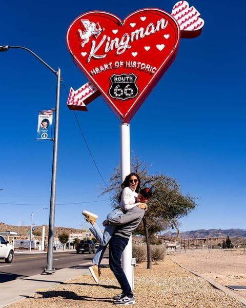 a man holding a woman in front of a heart shaped neon sign which reads Kingman Heart of Historic Route 66