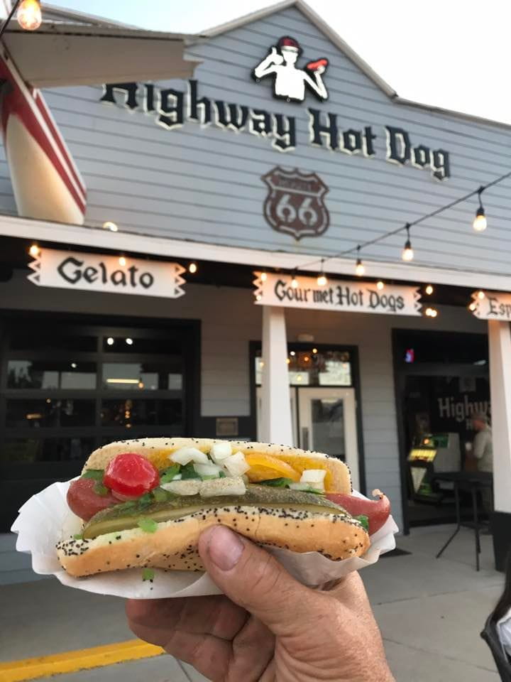 A hand holding a hot dog in front of the business Highway Hot Dog in Seligman, Arizona.