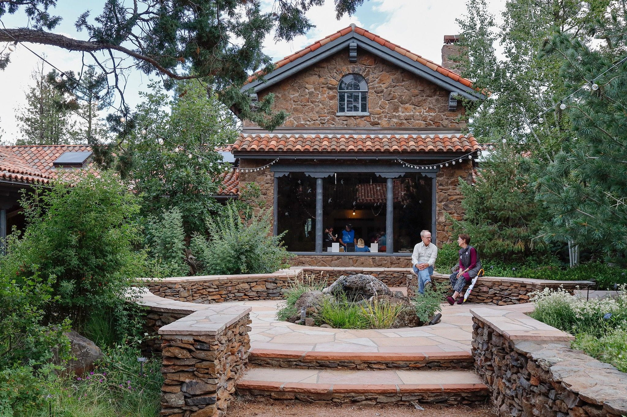 two people sitting on a short rock wall in front of a rock building surrounded by pine trees