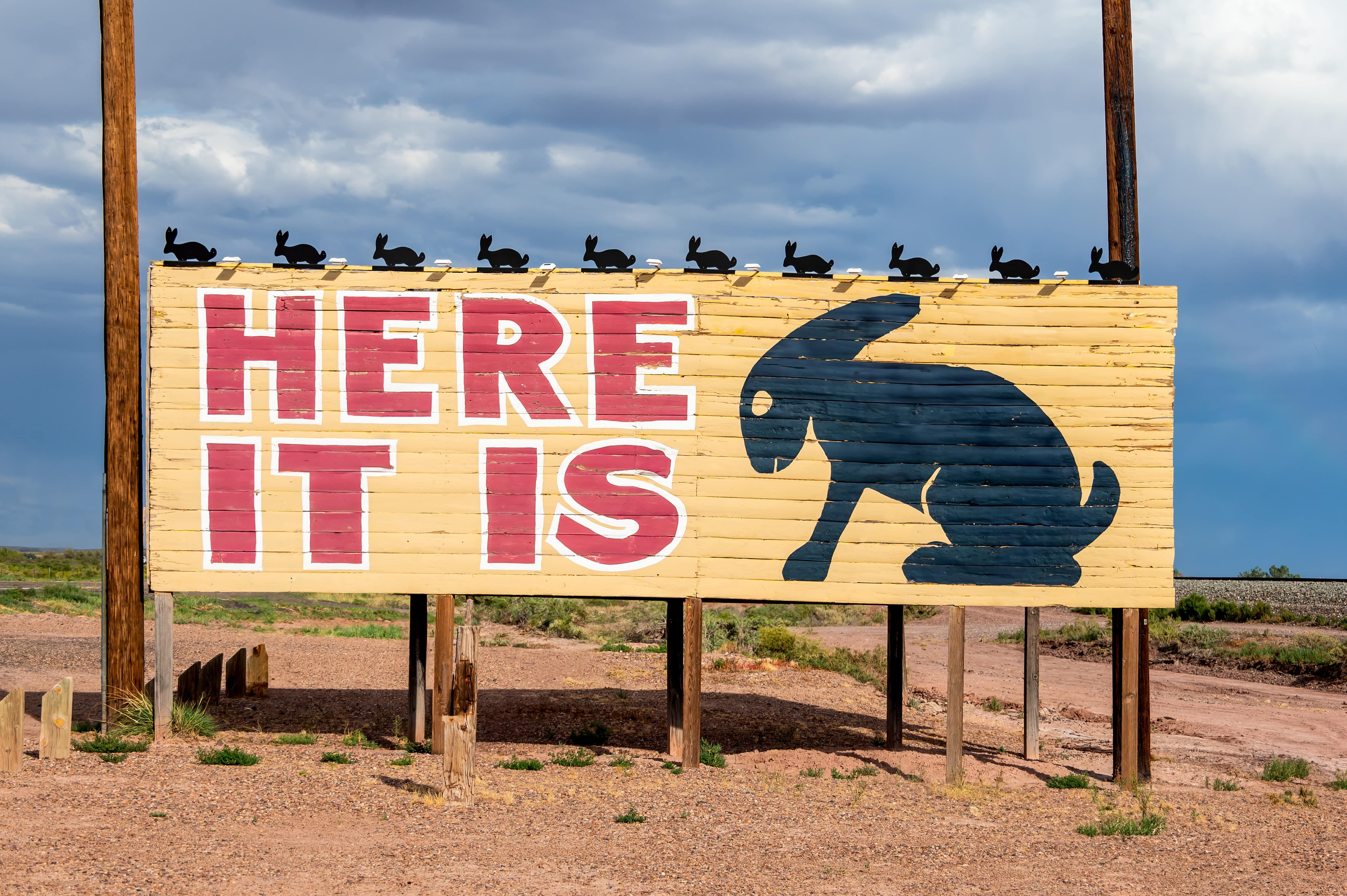yellow billboard with red letters that read HERE IT IS with a block silhouette of a rabbit