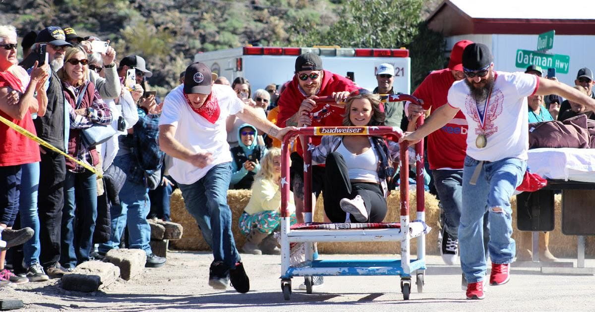 a crowd of people watch a group of 4 men run down the street while pushing a cart with a woman on top.