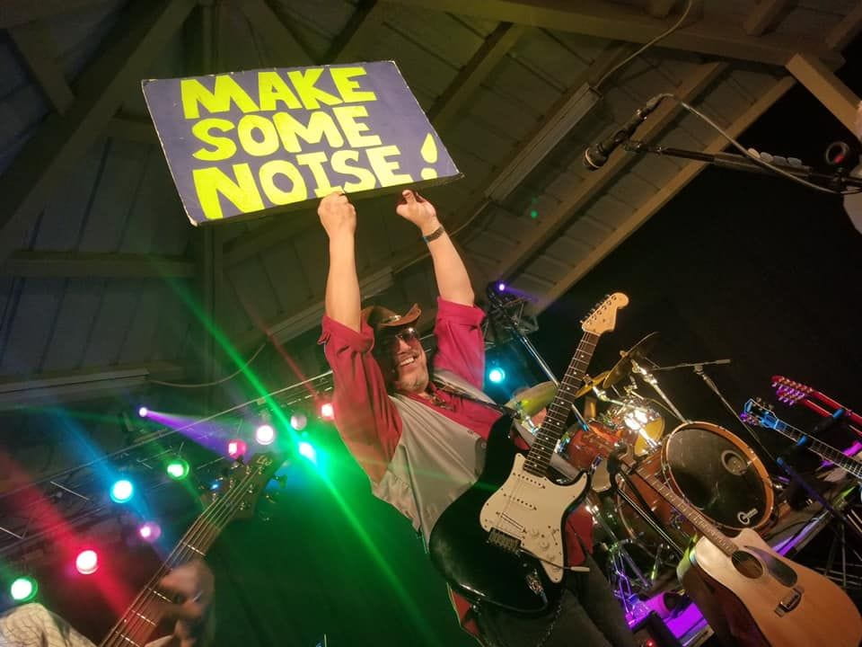 Man on a stage holding a sign that says, "Make some noise!"
