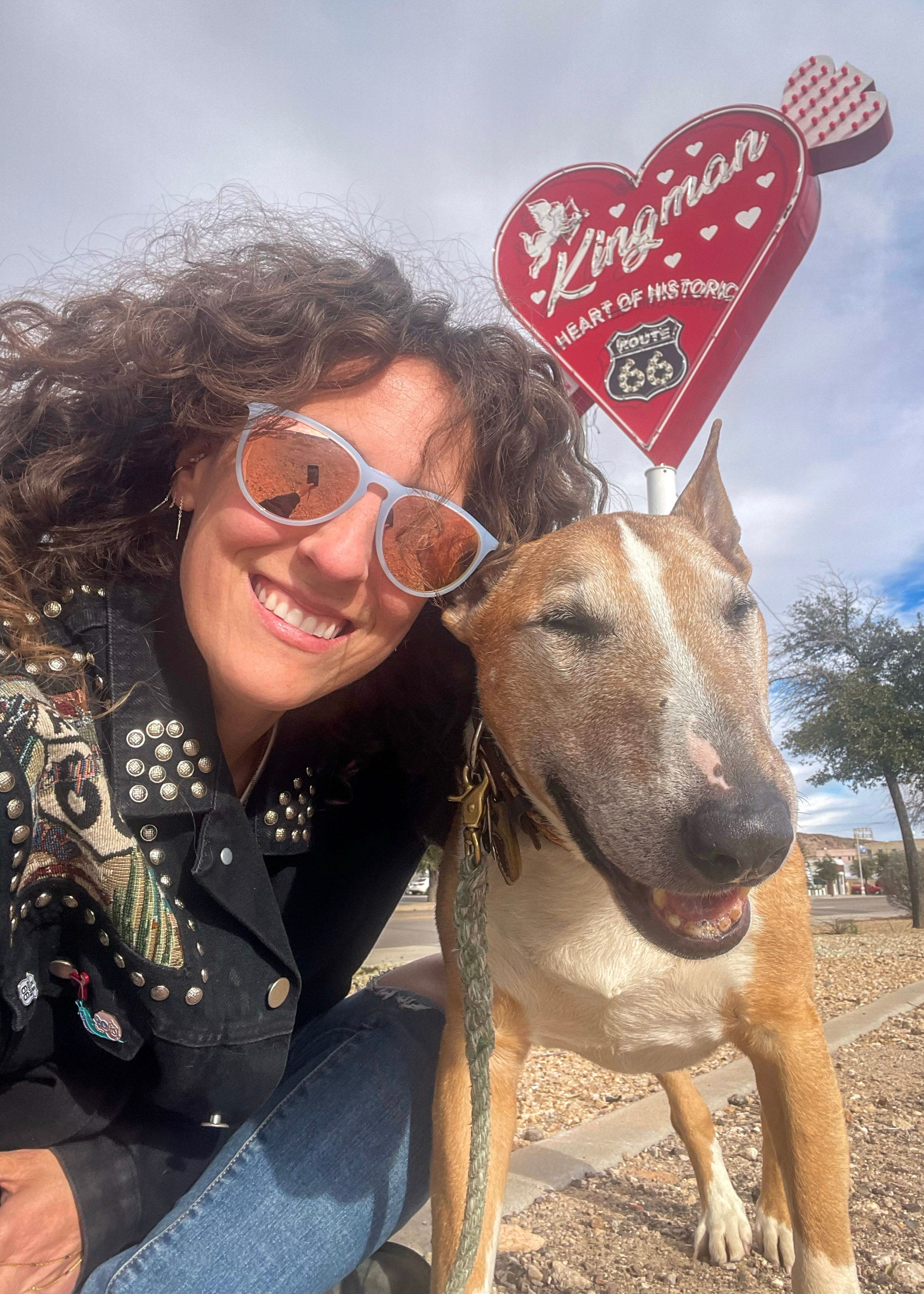 Woman wearing sunglasses poses with her pet dog in front of the Kingman heart neon sign